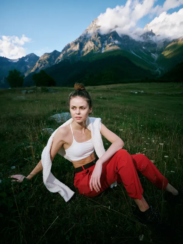 Mountains, landscape, outdoor, woman, meadow, grass, fashion, relaxed pose in a white crop top and red trousers resting on a lush meadow beneath blue sky and alpine peaks
