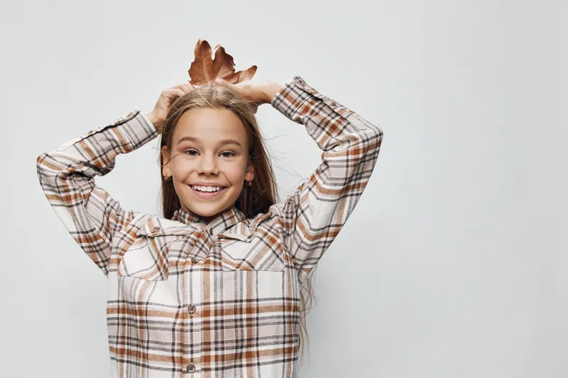 Girl with long hair and plaid shirt smiling and holding autumn leaves on head, happy child with natural light and neutral background for seasonal concept.