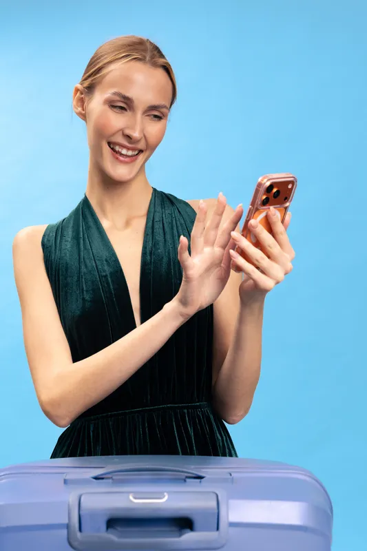 Smiling woman in black dress uses smartphone while standing behind purple suitcase on blue background. Concept of travel, technology, vacation planning, and modern lifestyle.