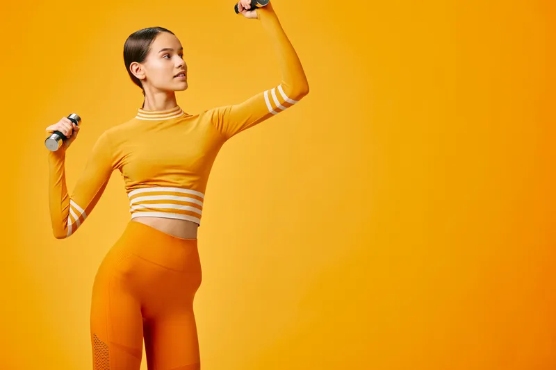 Athletic woman in orange fitness outfit lifting light dumbbells in studio. Monochrome bright orange background, confident upward pose, textured leggings and long sleeve crop top.