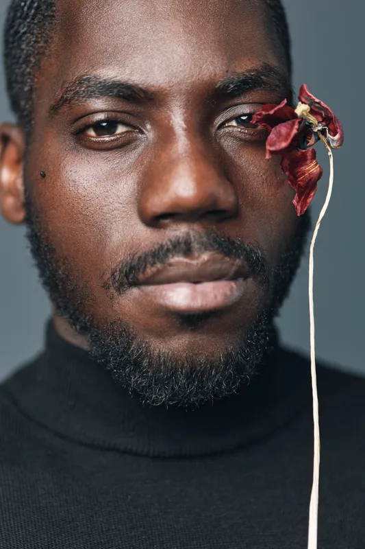 African man with dark skin and beard in creative conceptual style wearing black turtleneck with dried red flower near eye on gray background closeup portrait with serious expression.