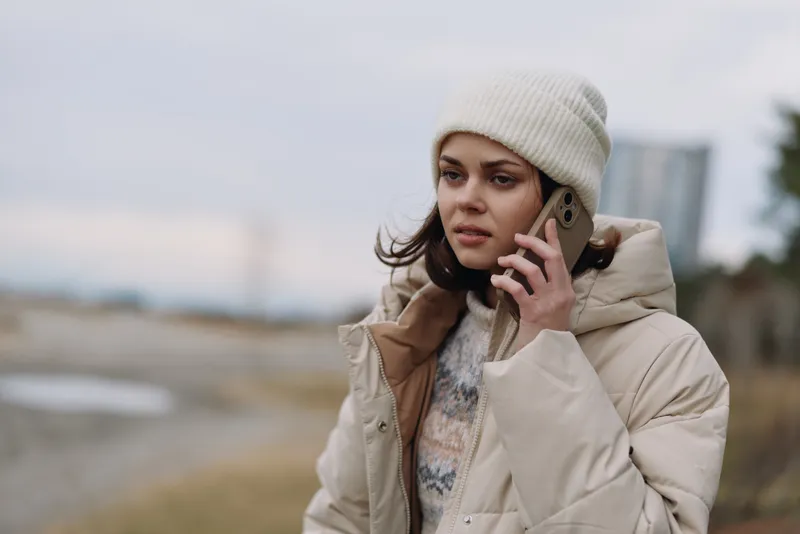 Woman in a beige puffer coat and knit hat stands outdoors, talking on a phone by the water in a cold winter setting, conveying communication and chilly mood.