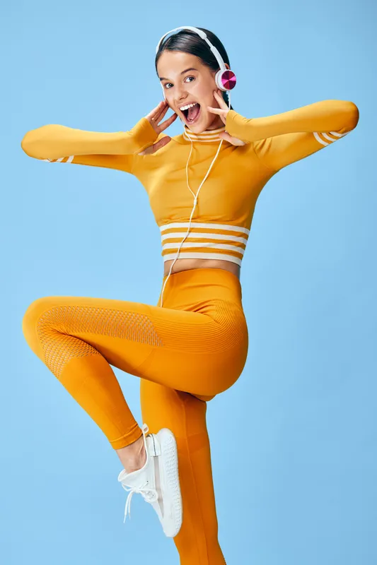 Energetic young woman fitness model in mustard yellow activewear and white sneakers poses on one leg against a solid light blue background, wearing headphones and smiling.