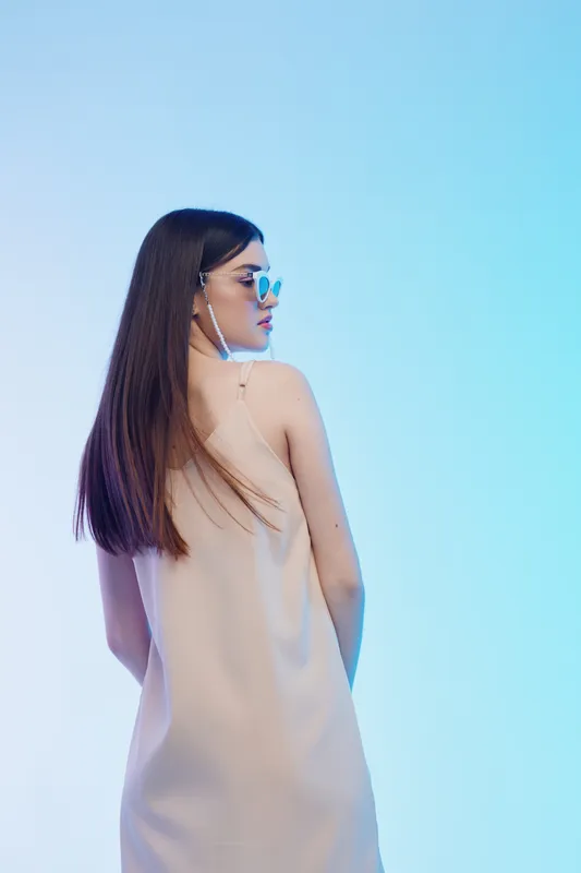 Young woman in a beige dress and sunglasses poses with her back turned against a blue gradient background. Concept of beauty, fashion, and modern style in studio lighting.