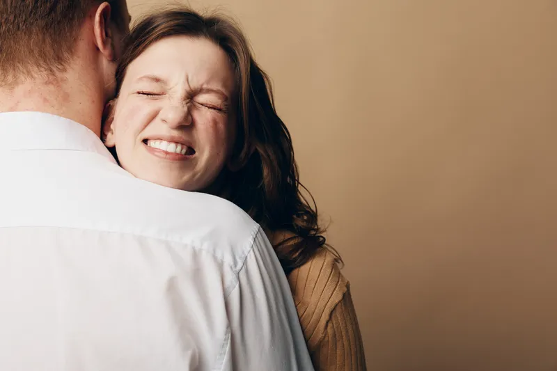 Happy woman with closed eyes embraces man wearing white shirt. Warm beige background creates cozy atmosphere. Concept of love, affection, relationship, and emotional connection between couple.