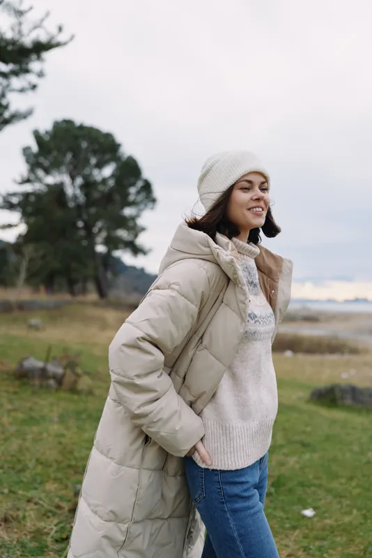 Woman in a warm winter coat and knitted sweater stands outdoors in a windy landscape, smiling softly as trees and open field create a calm casual fashion portrait.