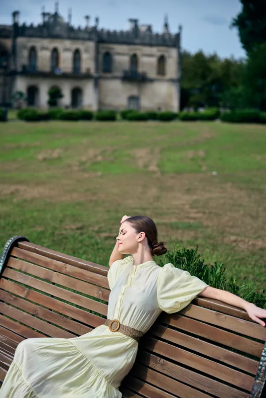 Woman in a pale vintage dress sits on a wooden bench in a sunny park, with a historic building in the background, radiating calm and timeless elegance.