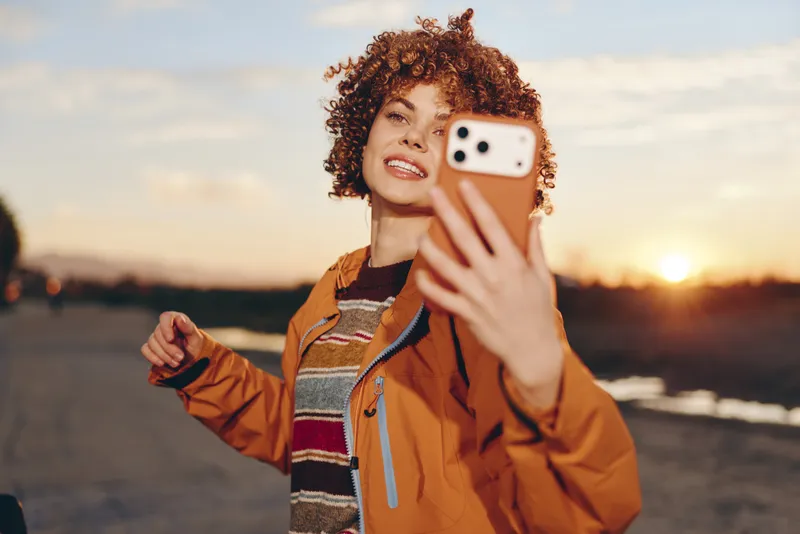Woman lifestyle portrait outdoors at sunset, curly hair and bright smile, orange jacket, holding smartphone for selfie, warm golden light on face, relaxed evening atmosphere by shoreline