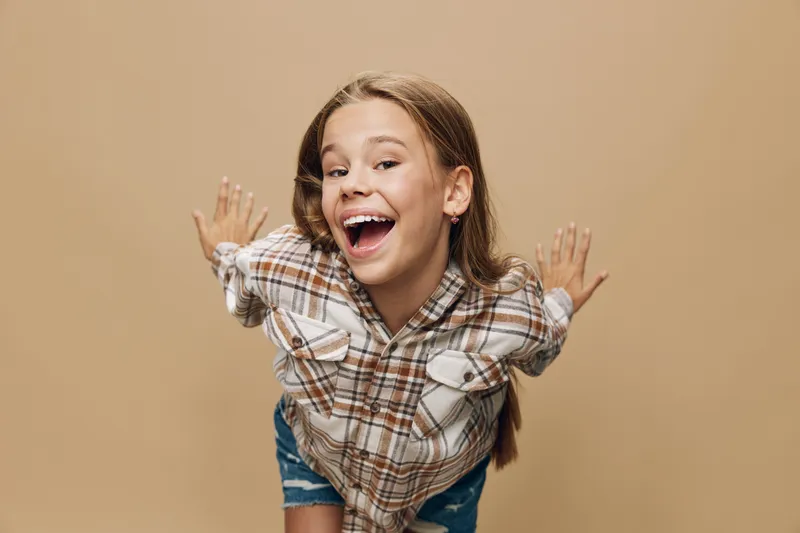 Happy girl smiling with hands spread out wearing plaid shirt and denim shorts standing against beige background for childhood and joy.