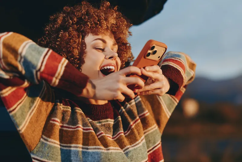 Woman with curly hair in striped sweater outdoors smiles while holding smartphone for selfie, golden hour light, scenic bokeh background, casual lifestyle moment, joyful expression today