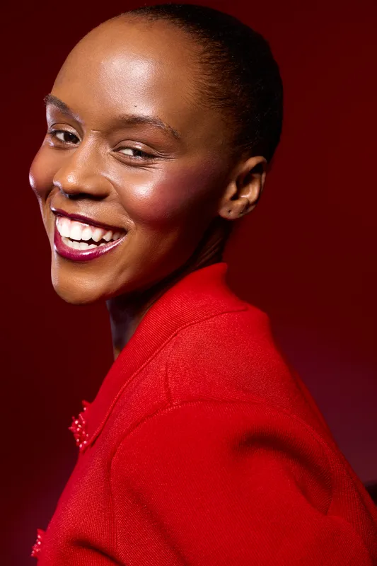 Smiling African American woman with dark skin wearing a red jacket poses against a red background. Studio portrait emphasizing beauty, confidence, and fashion with vibrant colors.