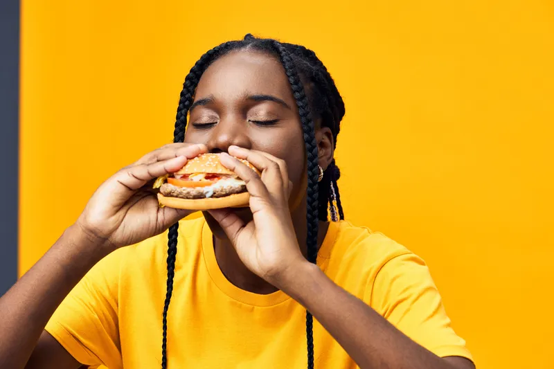 Young Black woman enjoying a delicious burger against a vibrant yellow background, conveying happiness and satisfaction with tasty fast food