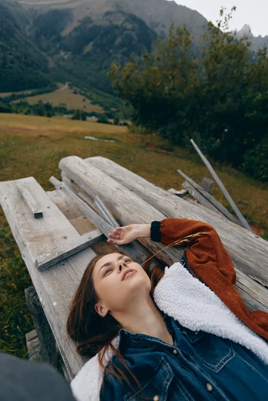 Woman portrait relax on wooden bench outdoors in countryside meadow with mountains and trees in background, casual jacket, serene expression and peaceful nature escape.