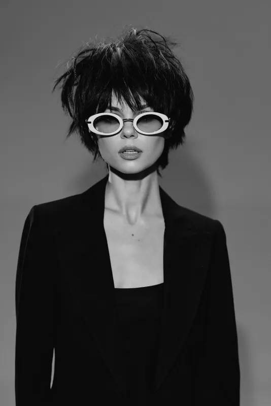 Black and white portrait of stylish woman wearing sunglasses and blazer with short hair in studio lighting.