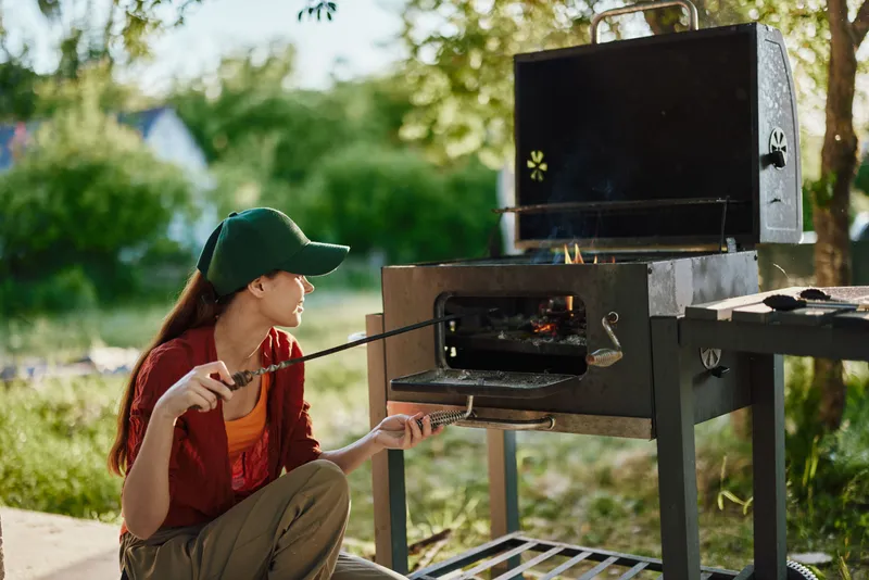 Young woman enjoying outdoor grilling in a sunny garden, wearing a green cap and casual clothes, engaged in a joyful cooking experience