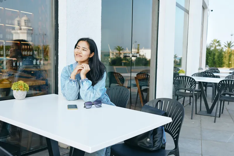 Thinking woman sits at an outdoor cafe table, sunlight and reflections, sunglasses on the table, notebook and phone nearby, modern terrace setting, casual style, relaxed mood, urban lifestyle shot