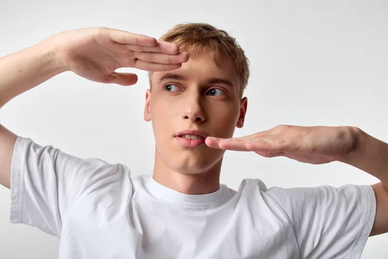 Man framing with hands wears a white shirt, creating a balanced pose in a bright studio, highlighting negative space and a focused expression for creative photographic framing