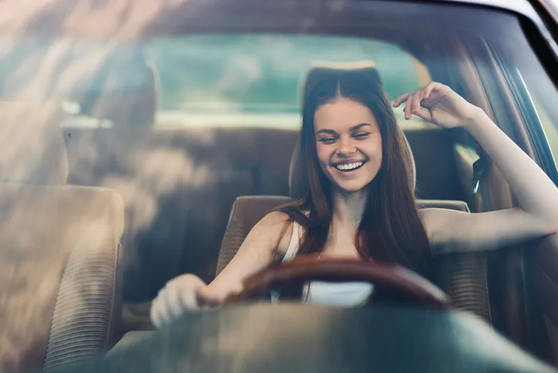 Happy young woman enjoying a carefree drive in a vintage car, wearing a casual outfit, radiating joy and freedom, with beautiful reflections in the glass.