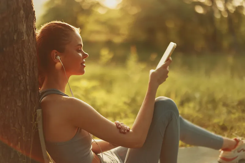 Young woman enjoying music while relaxing outdoors in a tranquil setting, wearing casual athleisure attire with headphones, against a warm, natural background
