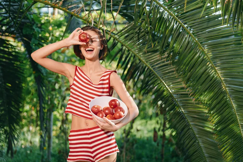 A cheerful person wearing a red and white striped outfit stands outdoors among lush greenery, smiling while holding a tomato up to their eye, radiating a playful, healthy vibe.