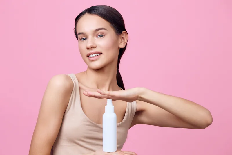 Young woman promoting a skincare product with a cheerful smile against a soft pink background, showcasing healthy skin and beauty concepts