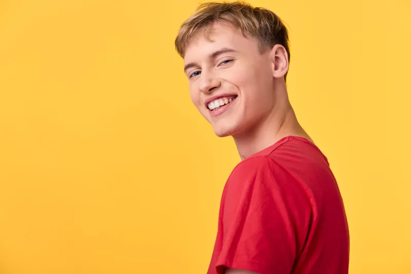 Smile, young man in a red shirt poses against a bright yellow background, showcasing happiness and confidence in a casual fashion shot with studio lighting and vibrant, approachable style