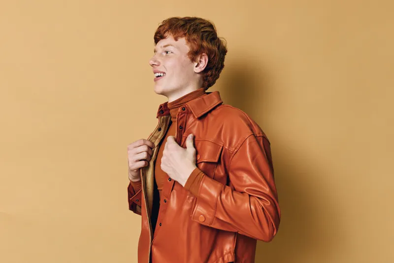 Young man with red hair wearing brown leather jacket standing and looking to side against beige background. Casual style, smiling expression and modern urban fashion portrait.