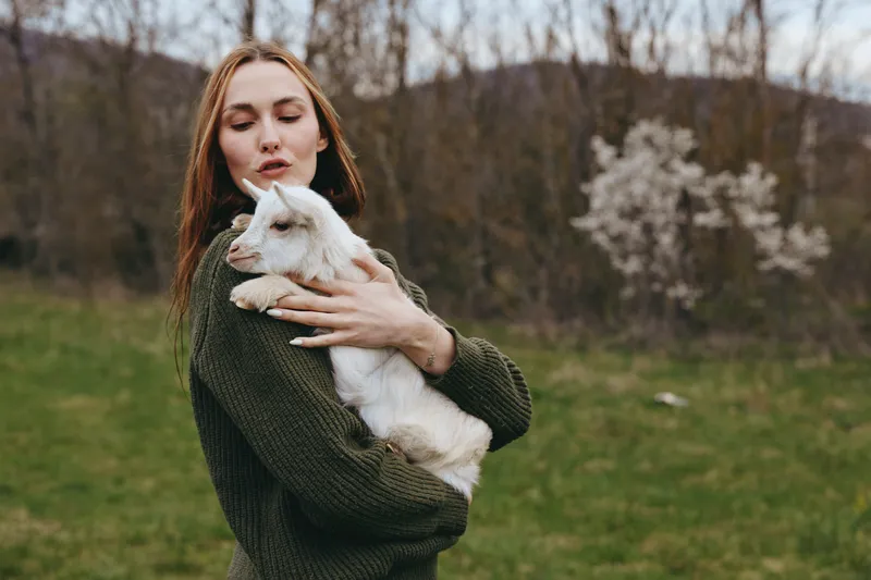 Caring woman holds a goat kid in an open meadow, outdoors under soft daylight. A lifestyle moment of friendship, affection, and nature, with a cozy, rustic mood and gentle bond.