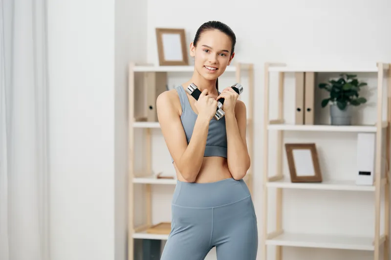 Woman fitness in athleisure stands with dumbbells in a bright home gym, showcasing workout routine, strength training, and focused energy for health and sport performance.