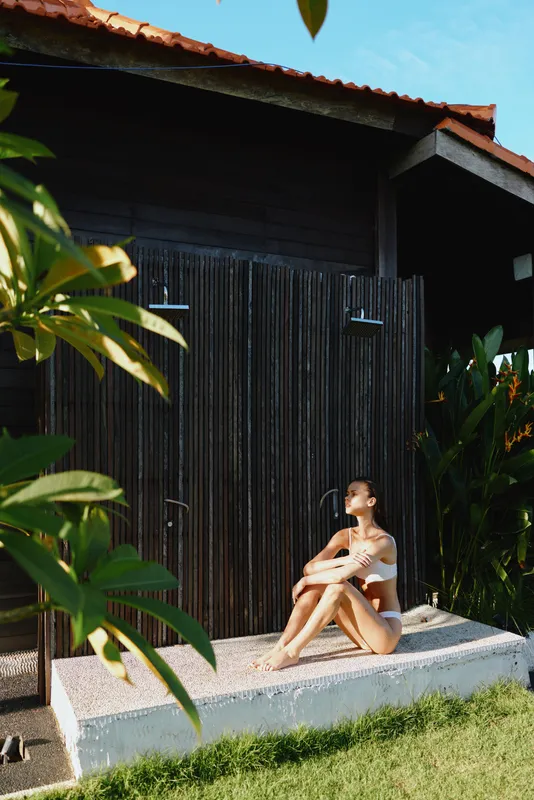 Young woman in white swimwear sitting on a stone ledge near a wooden building outdoors. Relaxed lifestyle concept with natural light and tropical greenery around her.