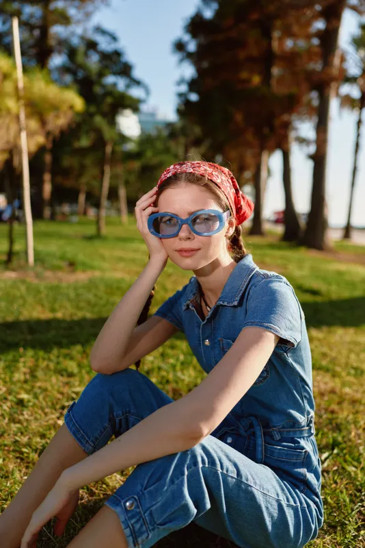 Lifestyle woman in modern boho-western denim street style wearing blue sunglasses and red bandana, sitting outdoors in warm sunlight with film color tones.