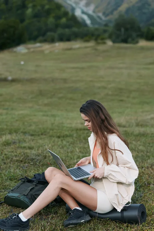 A focused outdoor professional sits on grass with a laptop, showcasing remote work in a natural setting. She types with intent, blending creativity and productivity in a calm environment.
