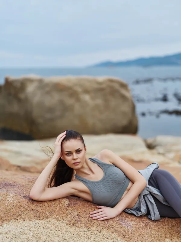 Fit young woman in athletic wear posing on rocky beach, expressing calmness and confidence against serene ocean backdrop, ideal for fitness and wellness themes