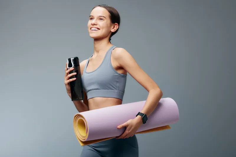 Athletic woman in a light blue sports bra and leggings, holding a water bottle and a rolled yoga mat, ready for a dynamic workout in a clean studio.