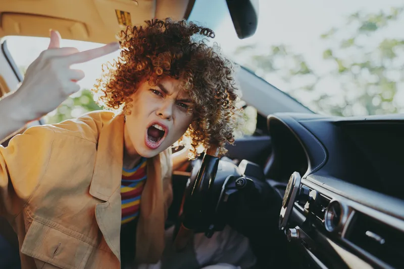 Young woman with curly hair sits in a car, shouting with open mouth and raised hand, capturing a bold, candid moment of frustration and energy
