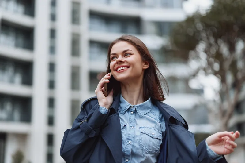 A smiling woman in a denim shirt and navy jacket talks on a phone outdoors in an urban setting, conveying casual style, friendly communication, and confidence.