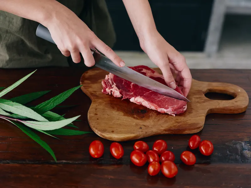 Женщина с ножом в руке нарезает свежее мясо для стейка на жарку на кухне