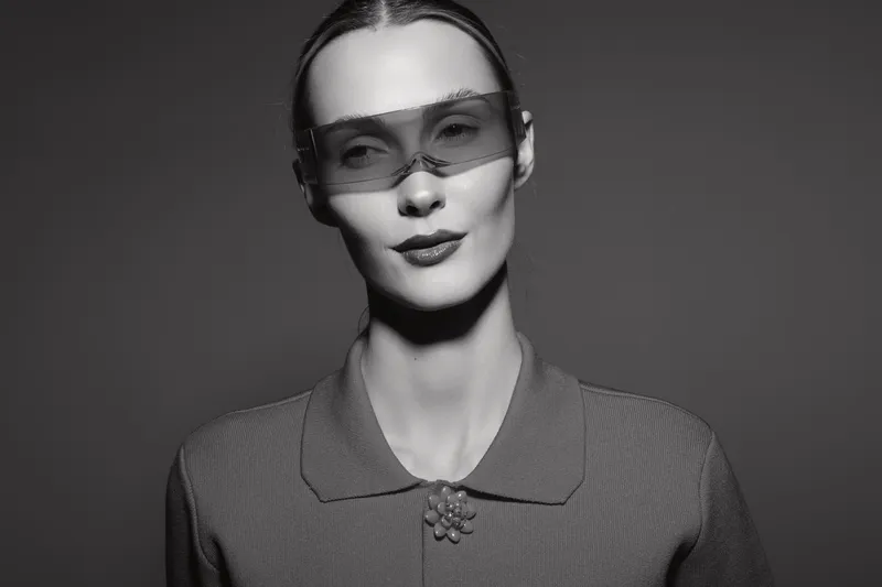 Portrait of a pale skinned white woman wearing translucent protective glasses and a collared shirt. Studio lighting creates a sharp and minimalist monochrome mood.