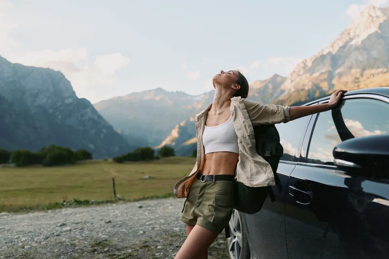 A confident woman leans against an open car door in a mountainous valley, basking in sunlit air and pine scent. A candid lifestyle moment of travel, exploration, independence, joy.