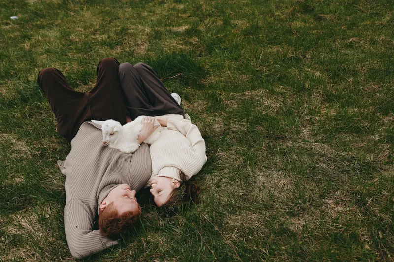 Lifestyle shot of a couple lying on grass embracing softly, with a playful goat kid nearby, capturing warm affection and natural connection in an outdoor rural setting.