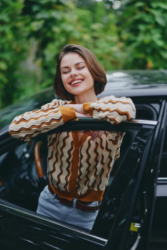 woman smiles happily as she leans on a car window, arms resting on the frame, radiating confidence and joy, a cheerful moment suggesting readiness for a pleasant journey