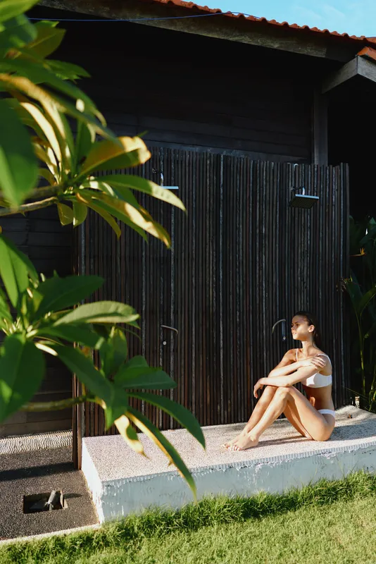 Relaxed woman sitting on a concrete step outside a wooden shed, wearing light swimwear. Outdoor lifestyle scene with greenery, sun, and a warm casual mood.