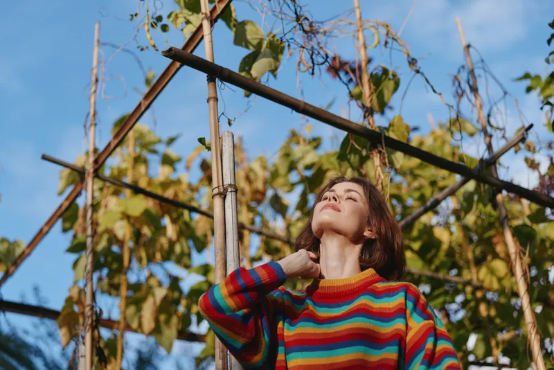 Woman in a colorful striped sweater, 20s-30s, standing in a garden among trellis and vines, looking up with closed eyes, relaxed and sunlit, lifestyle portrait for fashion and travel.