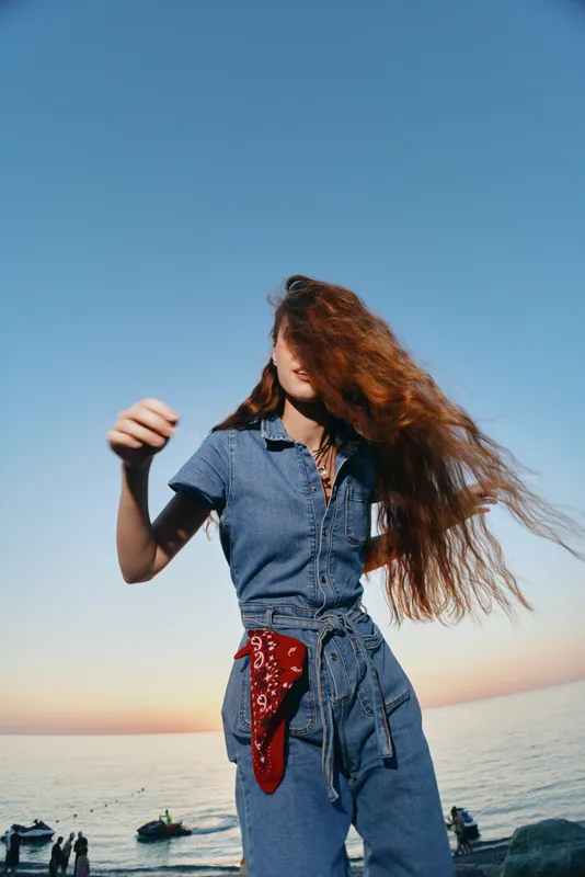 Lifestyle woman in modern boho-western denim street style stands by the sea with wind-blown red hair and warm film color tones