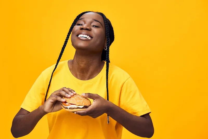 Happy young Black woman enjoying a delicious burger on a vibrant yellow background, celebrating food and good vibes with a joyful expression and casual style