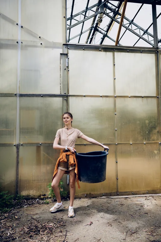 Woman gardening in greenhouse holding a large black bucket, young casual female in sneakers and shorts carrying container for soil and plants near glass structure