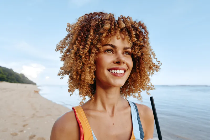 Woman with curly hair smiling on the beach near the ocean, outdoor summer portrait, soft sunlight and sea breeze, vacation travel mood, bright skin and happy expression, relaxed close pose