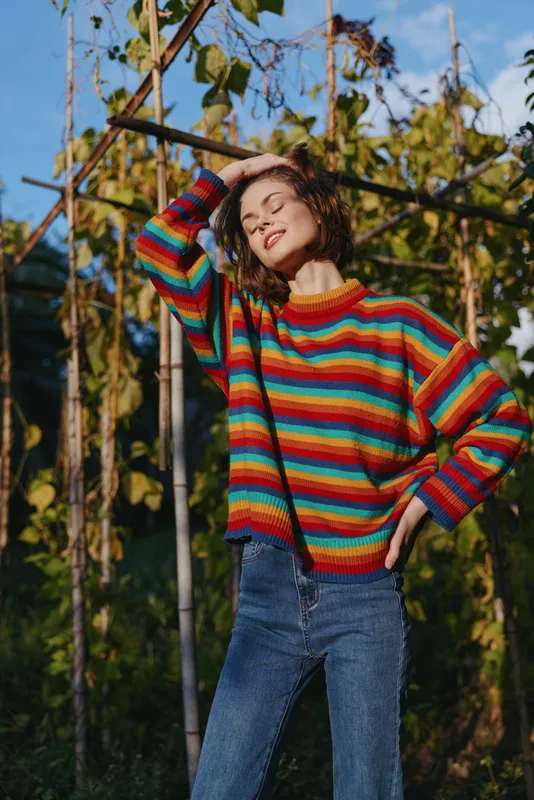 Woman in colorful sweater portrait outdoors wearing jeans, smiling with eyes closed and hand on head in sunlight in garden, casual fashion and relaxed mood.