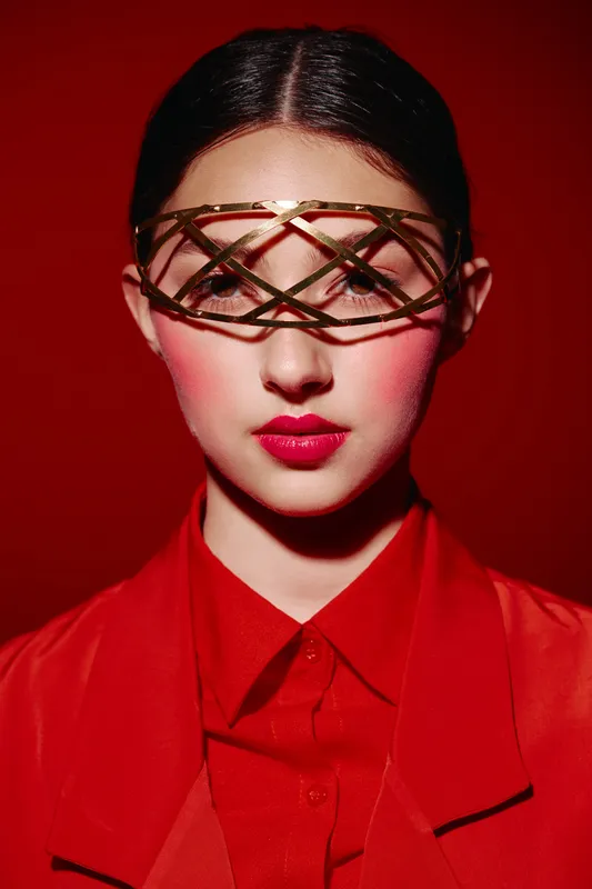 Young woman in dramatic red outfit wears gold futuristic accessory covering eyes. Studio portrait on red background. Concept of beauty creative fashion and avant-garde style.