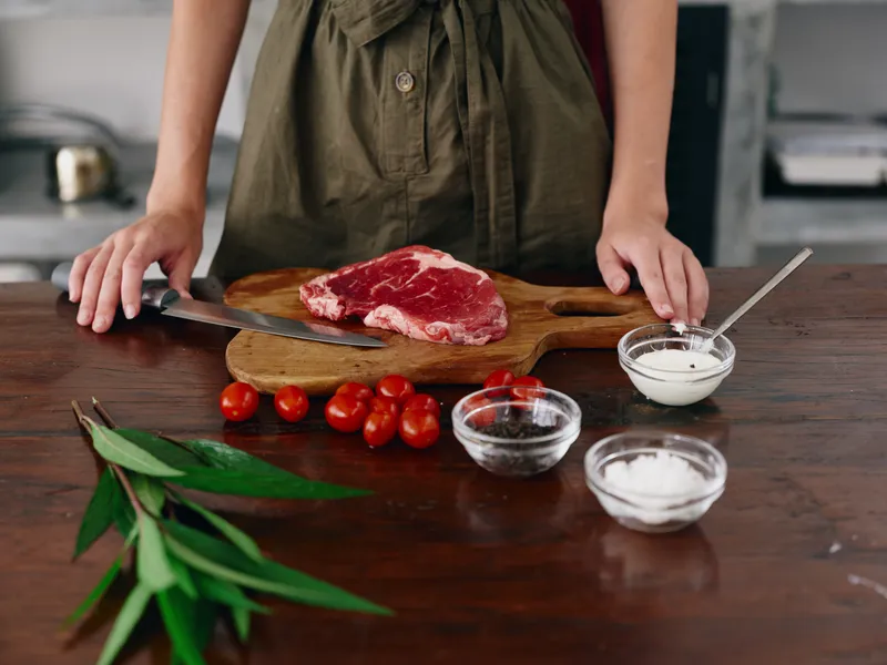 Женщина с ножом в руке нарезает мясо для стейка на жарку на кухне